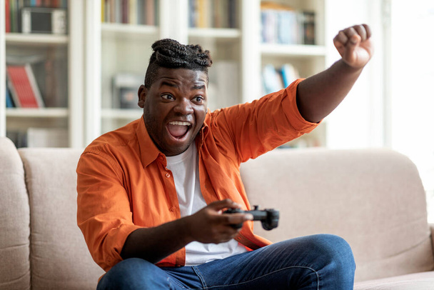 自宅で休憩、国内のエンターテイメントの概念。感情的な若いですアフリカ系アメリカ人男性とともにビデオゲームをしたり手を上げて笑ったりしながら一人でソファの上に家に座ってコピースペース - 写真・画像