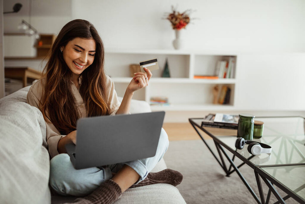 Smiling millennial mixed race woman shopaholic typing on laptop, using credit card, enjoy online shopping in living room interior. Business remotely, sale, banking and cashback at home - Photo, Image