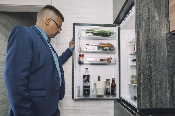 Mężczyzna szukający jedzenia w otwartej lodówce w kuchni późno w nocy. Wygląda jak głodny kawaler.. - Zdjęcie, obraz