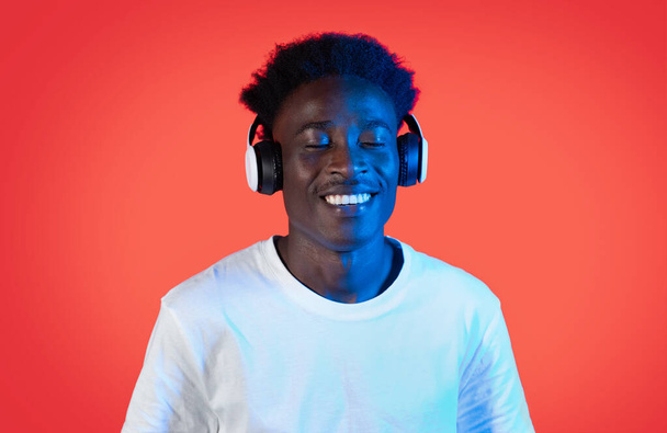 Fröhlich fröhlicher junger Schwarzer in weißem T-Shirt, der mit geschlossenen Augen Musik hört und lächelt, mit modernen drahtlosen Kopfhörern in Neonlicht, rotem Hintergrund, Nahaufnahme, Kopierraum - Foto, Bild