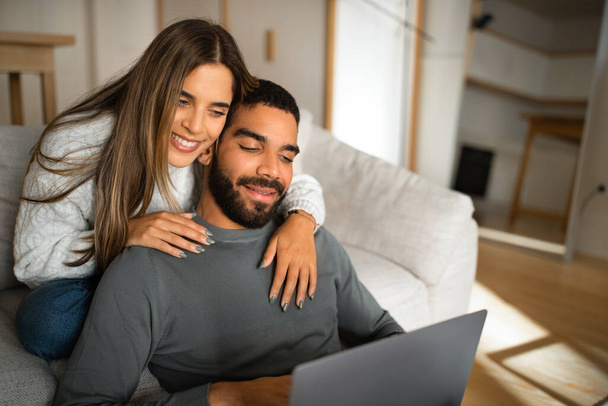 Cheerful multiethnic millennial couple sit on sofa, hugging, looking at laptop, have video call in living room interior. Love and relationship, communication remotely, new normal - Fotografie, Obrázek