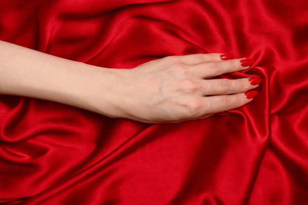 美しい赤マニキュア上の女性の手上の高級赤いシルクの背景.  - 写真・画像