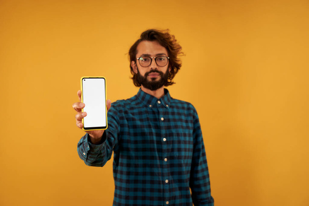 Young bearded hipster young man with glasses showing mobile screen in white isolated on yellow background. Boy in blue plaid shirt posing in studio portrait. Copy space. Serious guy holding cell phone - Foto, imagen