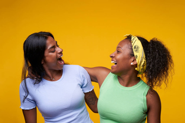 Zwei schöne schwarze Mädchen, die sich umarmen und komisch vereinzelt auf gelbem Hintergrund aussehen. Ein junges glückliches lesbisches Paar posiert überrascht in einem Studio. Völkerfreundschaft und homosexuelle Beziehung. - Foto, Bild