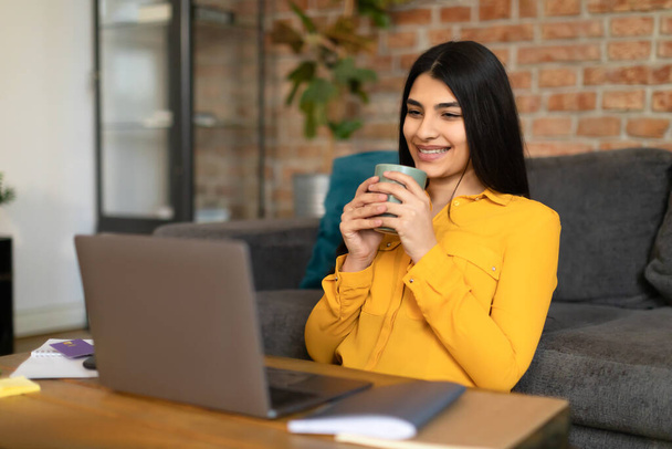 Positieve Spaanse dame die video 's bekijkt op internet terwijl ze thuis koffie drinkt, laptop gebruikt, naar het scherm kijkt en lacht, film kijkt en rust - Foto, afbeelding