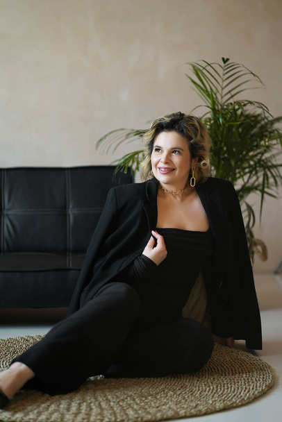 A smiling woman in a black suit is sitting on a rug near a sofa. - Фото, зображення
