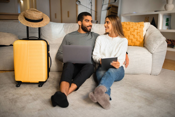 Smiling international millennial couple sit on floor with gadgets, suitcase and hat, choose country for vacation in living room interior. Relationships and love at home, travel planning app - Photo, Image