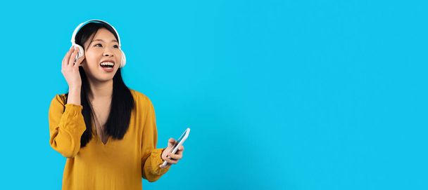 Joyful happy millennial asian woman in casual listening to music, using modern wireless stereo headphones and smartphone, looking at copy space, blue studio background, entertaining musical app - Fotoğraf, Görsel