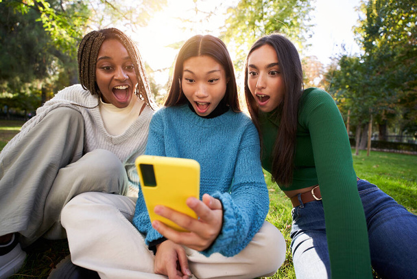 Three young women using a cell phone in the park. Portrait of teenage girls surprised looking at social networks on mobile. People watching the telephone screen fascinated with their mouths open. - Fotografie, Obrázek