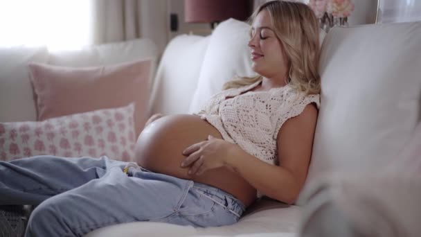Happy and relaxed young pregnant mother touching her belly sitting on sofa at home. Smiling Caucasian woman at the end of pregnancy waiting for the birth of her baby. People inside the house. - Footage, Video