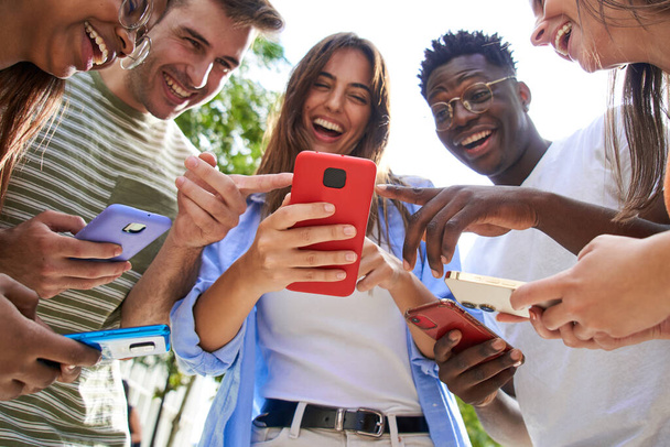 Niedrigwinkel-Ansicht der Gruppe lächelnde multirassische junge Studenten mit Handy, die lustige Rollen auf dem Handy beobachten. Versammelt lachende Menschen, die auf Telefone schauen und darauf zeigen. Soziale Beziehungen und Freundschaft im Freien.  - Foto, Bild