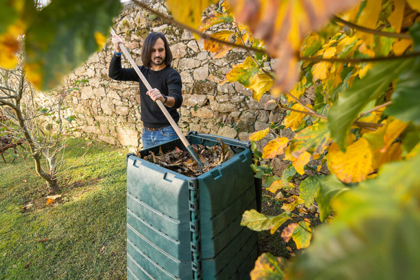 Um homem está misturando o lixo orgânico com folhas secas em um caixote do composto ao ar livre colocado em um jardim para reciclar resíduos domésticos e de jardim. Conceito de reciclagem e sustentabilidade - Foto, Imagem