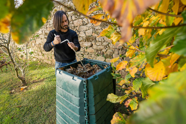 Мужчина смешивает органические отходы с сухими листьями в открытом компостном контейнере, помещенном в сад для переработки бытовых и садовых отходов. Концепция переработки и устойчивого развития - Фото, изображение