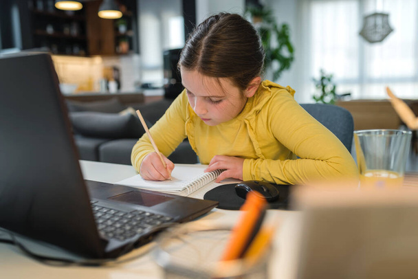 オンライン学習のためのラップトップコンピュータを使用してヘッドフォンでかわいい子供。ホームスクール。女の子は家で宿題をしている。家庭教育のためのライフスタイルコンセプト。社会的距離と教育. - 写真・画像
