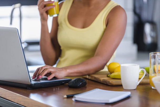 Junge fröhliche Frau surft zu Hause in der Küche auf einem Laptop im Internet und erfrischt sich mit einem Fruchtsaftgetränk - Foto, Bild