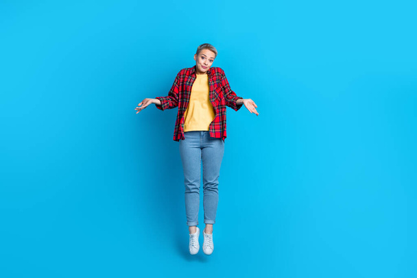Full length photo of sporty positive woman wear checkered shirt jeans shrugging shoulders jumping isolated on blue color background. - Foto, Imagen