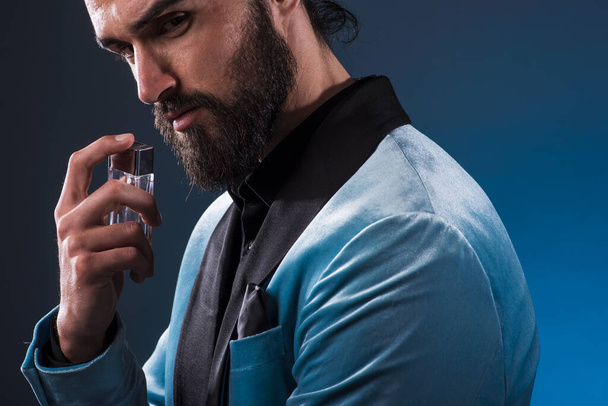 Beautiful masculine bearded young man holding a bottle of fragrance. Masculine perfume, bearded man in a suit. - Photo, image