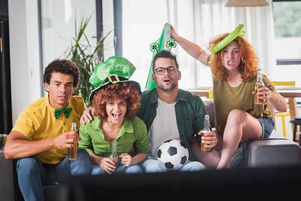 Πολυεθνική ομάδα νέων που διασκεδάζουν, βλέποντας ένα ποδοσφαιρικό παιχνίδι στην τηλεόραση. Επευφημίες για την Ιρλανδία. - Φωτογραφία, εικόνα