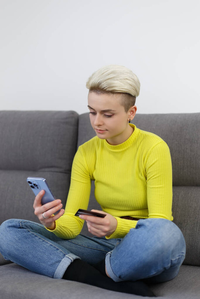 Beautiful white woman with short hair sitting on a couch at home and making a payment with smart phone and credit card - Photo, Image