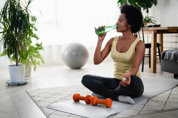 Afroamerikanerin macht zu Hause Übungen auf dem Boden, macht eine Pause und trinkt Wasser. Hydratation. - Foto, Bild