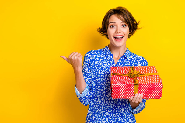 Photo of impressed funny woman dressed blue blouse holding red present box thumb empty space isolated yellow color background. - Foto, Bild