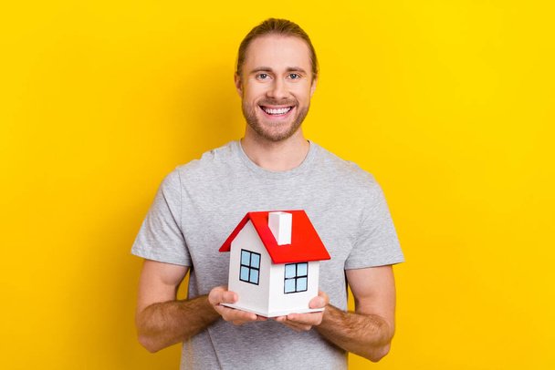 Photo of cheerful candid man wear grey stylish clothes hands hold mini house showing his dream isolated on yellow color background. - Fotografie, Obrázek