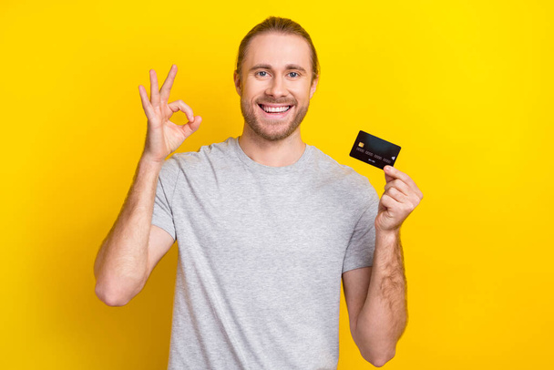 Photo of positive nice bearded man wear stylish grey clothes hold plastic card demonstrate okey symbol isolated on yellow color background. - Fotoğraf, Görsel