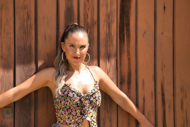 Young and beautiful woman, with ponytail, wearing a floral top and jeans, serious and angry with open arms leaning on a wooden door. Concept beauty, seriousness, anger, reproach. - Photo, Image