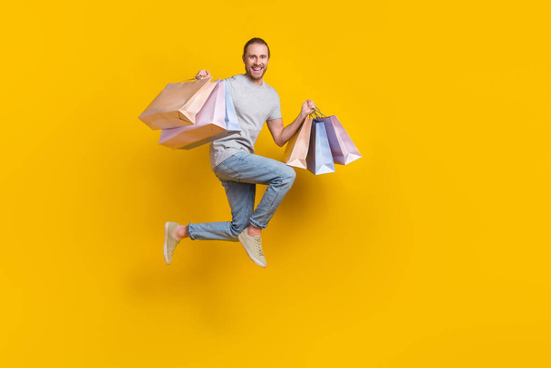 Full length photo of funky positive guy dressed grey t-shirt jumping high rising bargains empty space isolated yellow color background. - Foto, immagini