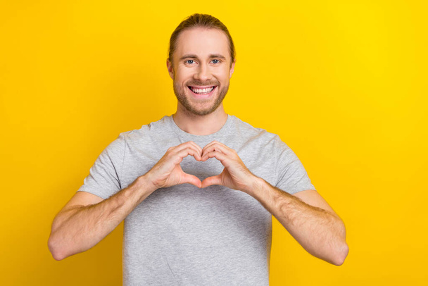 Photo of cheerful funny man wear grey t-shirt smiling showing fingers heart isolated yellow color background. - Photo, image