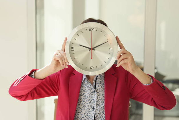 Woman covers face with big round wall clock standing in office. Employee in red jacket holds clock with hands. Time management at work - Foto, Bild