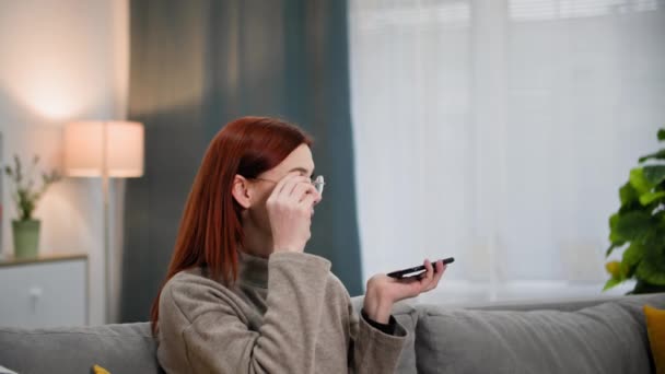 νεαρή γυναίκα σε γυαλιά για την όραση μιλάμε στο κινητό τηλέφωνο σε ηχείο παραγγελία παράδοση των ειδών παντοπωλείου ή σε απευθείας σύνδεση, ενώ χαλαρώνετε στο σπίτι - Πλάνα, βίντεο