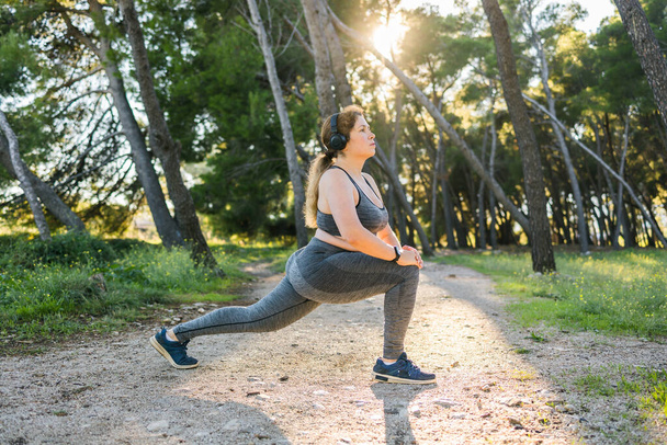 Adorable fat woman in tracksuit is engaged in fitness outdoor side view portrait. Young overweight woman lunges outdoors on warm summer day. Healthy lifestyle and weight loss. - Photo, Image