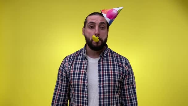 Droevige volwassen man in feestelijke feestmuts die verjaardagsfeestclaxon blaast, met opgeblazen ballon geïsoleerd op gele achtergrond. Overstuur mannelijke persoon viert vakantie alleen. Saaie boel. Eenzaamheid concept - Video