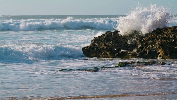 Güneşli bir sabahta kayaları vuran beyaz dalgalar. Fırtınalı okyanus manzarası. Yazın kumlu sahil şeridinde aşırı köpüklü deniz akıntısı. Büyük muhteşem su akıntısı dalgalanan kıyıya vuruyor.. - Fotoğraf, Görsel