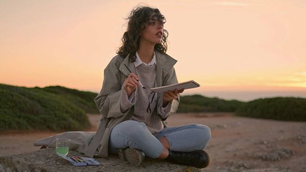 Young woman painting sunset mountain. Focused talented student admiring nature landscape resting on rock. Inspired beautiful artist girl drawing in sketch book at evening sky. Artwork leisure concept. - Photo, image