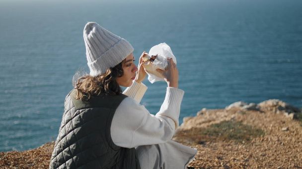 Podróżniczka jedząca kanapki na skalistym urwisku. Turysta zrobić sobie przerwę na wycieczkę pieszą korzystających śniadanie na spokojny widok na ocean. Samotna młoda kobieta odpoczywająca nad morzem w wiosenny weekend. Koncepcja pikniku na świeżym powietrzu - Zdjęcie, obraz