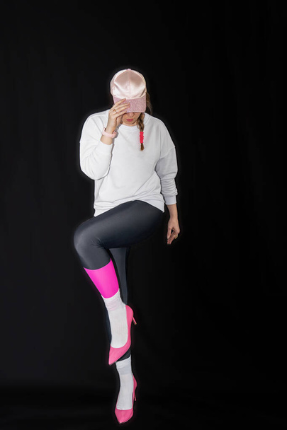 jonge vrouw draagt trendy kleding geïsoleerd op een zwarte achtergrond. Stijlvol model in wit sweatshirt, zwarte legging, witte sokken en roze hakken schoenen, met roze kap poseert voor de camera, volledige lichaamslengte  - Foto, afbeelding