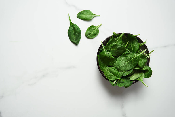 A bunch of Fresh baby spinach leaves in the in a black ceramic bowl on white marble background. Top view and flat lay photo with copy space. High quality photo - Photo, Image