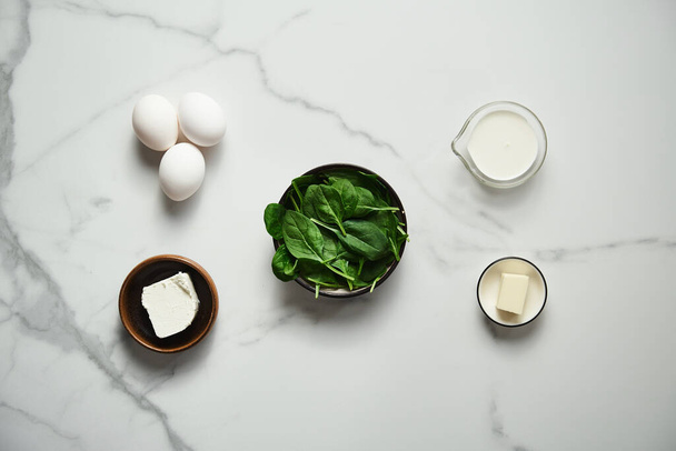 Iingredients for healthy spinach and feta omelette. Eggs with heavy cream, Fresh baby spinach leaves in the black ceramic bowl, white marble background. High quality photo - Foto, Imagem