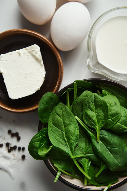 Iingredients for healthy spinach and feta omelette. Eggs with heavy cream, Fresh baby spinach leaves in the black ceramic bowl, white marble background. High quality photo - Photo, Image