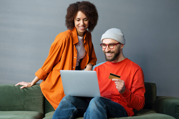 Schöner kaukasischer bärtiger Mann in lässiger Kleidung und Brille, der mit seiner Freundin auf einem Sofa sitzt, Kreditkarte in der Hand, Laptop benutzt, per Internet-Banking bezahlt, online einkauft - Foto, Bild