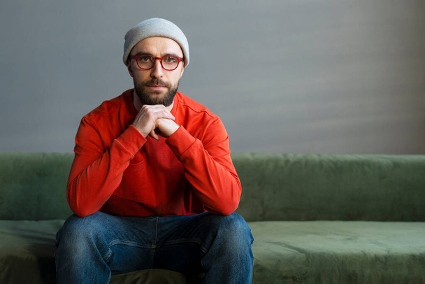 Millennial blanke knappe bebaarde man, het dragen van trendy bril, zitten op de bank op het werk of loft appartement, kijken naar de camera met inzichtelijke blik. Beroepsberoep, freelance portret - Foto, afbeelding