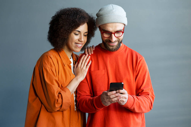 Afroamerykanka młoda ładna kobieta stoi blisko swojego stylowego chłopaka za pomocą telefonu komórkowego, przewijanie wiadomości, sprawdzanie treści w mediach społecznościowych, SMS-y online wiadomości, na tle szarej ściany - Zdjęcie, obraz