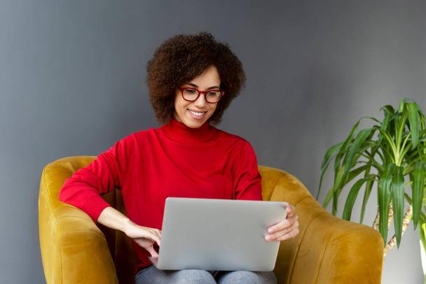 Joven mujer afroamericana gerente ejecutivo, mujer de negocios en línea de trabajo, escribiendo texto en el ordenador portátil, sentado en un sillón cómodo en una oficina corporativa contemporánea. Negocios. Gente. Tecnología - Foto, Imagen
