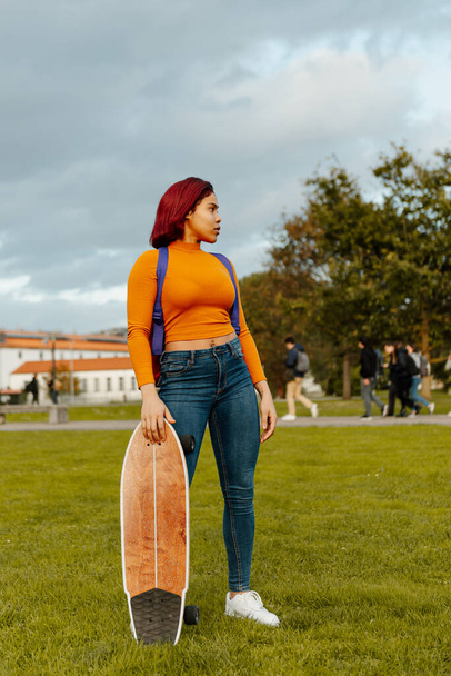 Portret nastoletniej Latynoski stojącej w plecaku, dżinsach i pomarańczowej bluzie, trzymającej deskorolkę przed kampusem uniwersyteckim. kultura młodzieży i powrót do szkoły - Zdjęcie, obraz
