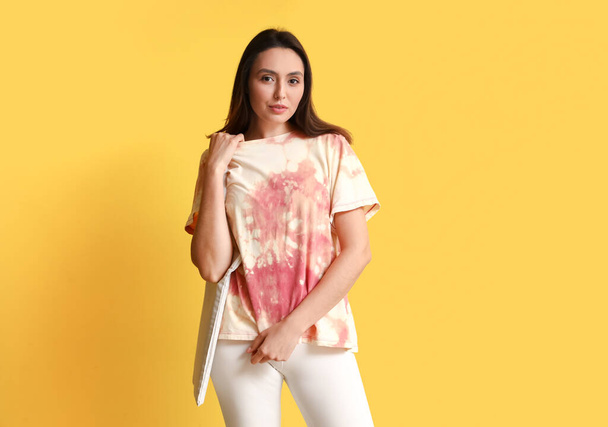 Stylish young woman in tie-dye t-shirt and with bag on yellow background - Фото, изображение