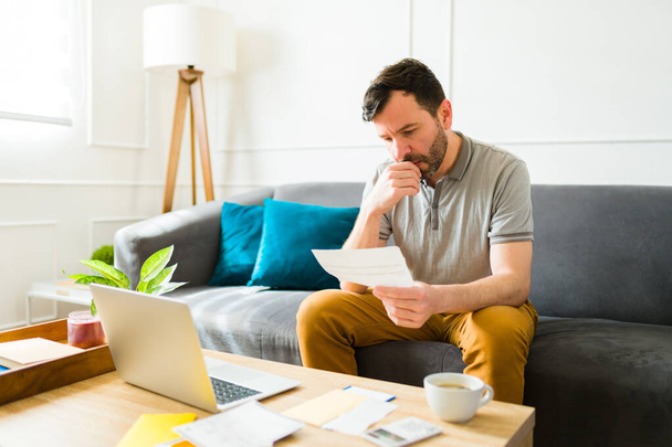 Skupiony biały człowiek patrząc na swoje finanse i robiąc swoje podatki za pomocą laptopa siedzi na kanapie w salonie - Zdjęcie, obraz