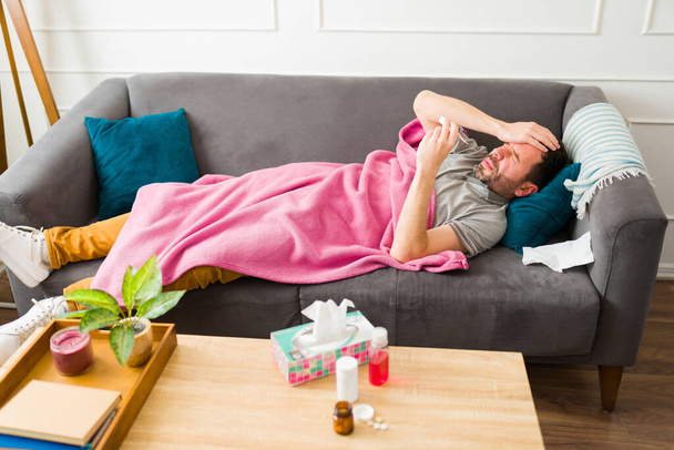 Verärgerter kranker Mann sieht müde aus, während er in eine Decke gehüllt auf dem Sofa liegt und Fieber und Erkältung bekommt - Foto, Bild