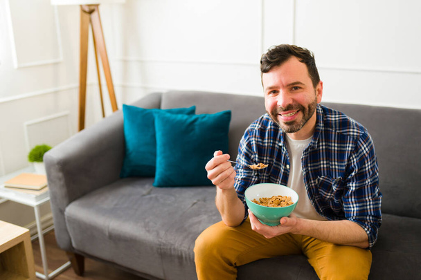 Όμορφος, ευτυχισμένος άντρας γύρω στα 30, τρώει δημητριακά κάθεται στον καναπέ και απολαμβάνει το πρωινό του στο σαλόνι. - Φωτογραφία, εικόνα